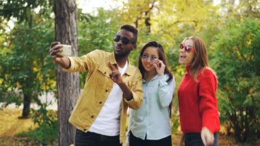 Şık genç insanlar adam ve kadın poz ve parkta yürüyüş sırasında holding smartphone gülümseyen güneş gözlüğü takmış selfie alıyor. İnsan ve teknoloji kavramı.