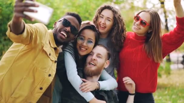 Elegantes jóvenes niñas y chicos están utilizando el teléfono inteligente para tomar selfie en el parque posando para la cámara y riendo. Los estudiantes están usando ropa casual y gafas de sol . — Vídeo de stock