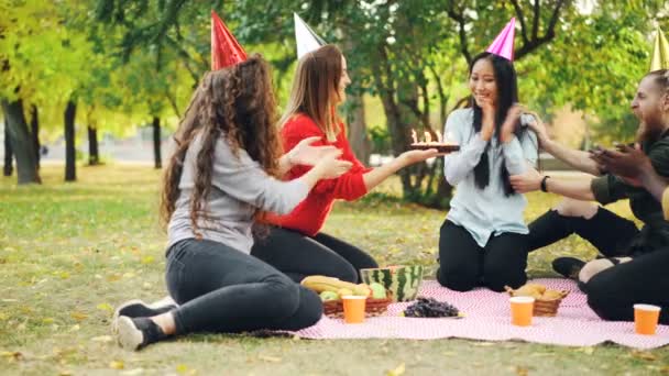 Amigos están felicitando a mujer asiática en cumpleaños dando pastel haciendo sorpresa, chica está soplando velas, sonriendo y aplaudiendo las manos durante la fiesta al aire libre en el parque . — Vídeos de Stock