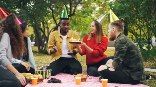 Bearded African American guy is having birthday party in park blowing candles on cake and laughing enjoying surprise, his friends are clapping hands. — Stock Video