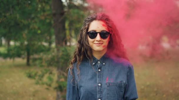 Slow motion portret van aantrekkelijke jonge vrouw in zonnebril permanent buitenshuis en camera te kijken terwijl gekleurde poeder gulal is haar bekogeld op Holi festival. — Stockvideo