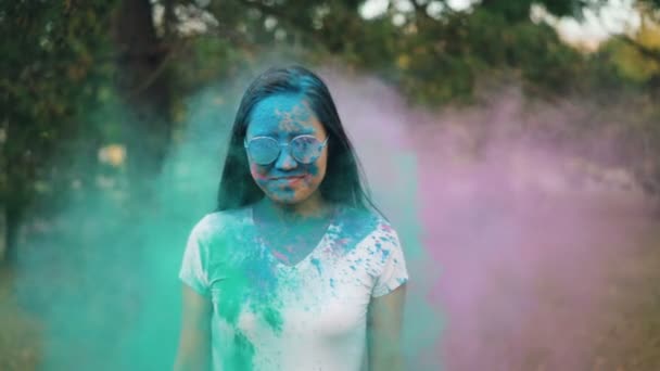 Retrato de una hermosa joven en gafas de sol disfrutando del festival Holi de pie al aire libre mientras la gente le arroja polvo colorido. Concepto de cultura y tradiciones . — Vídeo de stock