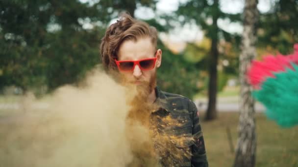 Retrato en cámara lenta de un hombre guapo con gafas de sol brillantes parado solo en el festival Holi con la cara recta y luego sonriendo y riendo cuando la gente le lanza pintura . — Vídeo de stock