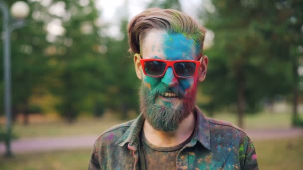 Treg film portrett av kjekk fyr på Holi festivalen med malt ansikt og trendy solbriller ser på kamera og smiler i parken alene . – stockvideo