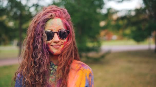 Zpomalený pohyb portrét šťastná dívka s dlouhými kudrnatými vlasy pokryté vícebarevná barva prášku na Holi festival stojí venku a usmívá se nosí sluneční brýle. — Stock video
