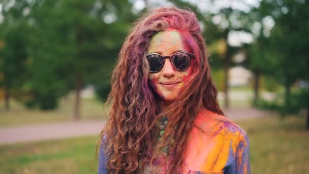 Retrato de una hermosa chica con gafas de sol de pie al aire libre con la cara y el pelo cubierto con pintura gulal de colores brillantes en Holi vacaciones. Concepto de juventud y tradiciones . — Vídeos de Stock