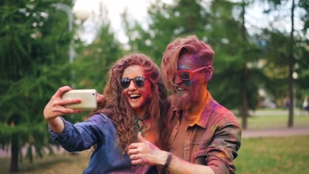 Chica y chico con caras y cabello cubierto de pintura se están tomando selfie junto con el teléfono inteligente de pie al aire libre en el parque y divertirse disfrutando de la fiesta y la tecnología . — Vídeos de Stock
