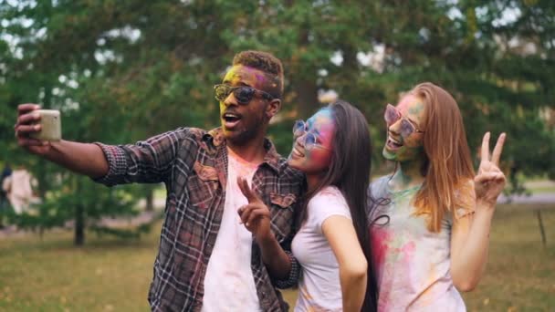 Rallentatore di uomo afroamericano in occhiali da sole scattare selfie con belle ragazze al festival di Holi, i loro volti, capelli e vestiti sono coperti di vernice . — Video Stock