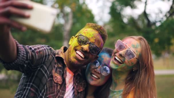 African American guy wearing sunglasses is taking selfie with female friends at Holi color holiday, their faces and hair are dirty with multicolor paint. — Stock Video