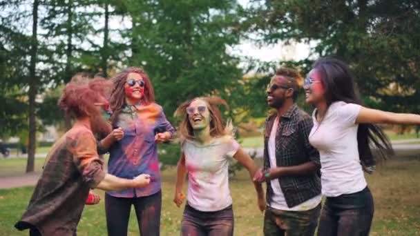 Slow motion of happy young people jumping then throwing powder paing at Holi festival outdoors in park. Faces, hair and clothes of students is colorful. — Stock Video