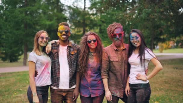 Retrato em câmera lenta de demônios sorridentes meninas e caras com rostos coloridos e roupas em pé ao ar livre e olhando para a câmera na festa. Holi festival e conceito de juventude . — Vídeo de Stock