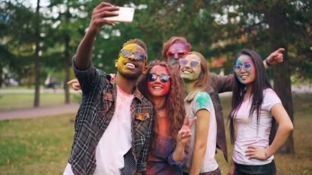 Zwolnionym tempie studenci happy wieloetnicznym grupy z kolorowe twarze i włosy biorąc selfie w parku przy użyciu aparatu w smartfonie i zabawy festiwalu Holi. — Wideo stockowe