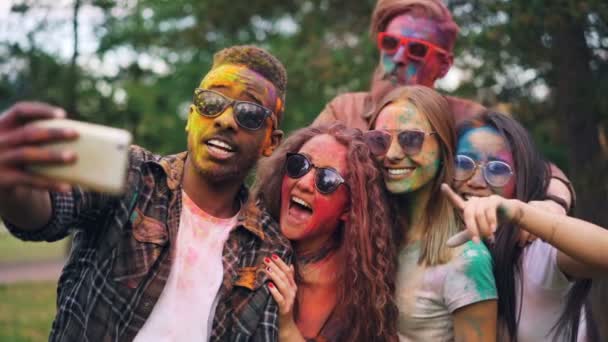 Excited friends girls and guys are taking selfie with colored faces and hair using smartphone, stylish young man is jumping and laughing. Modern technology and party concept. — Stock Video