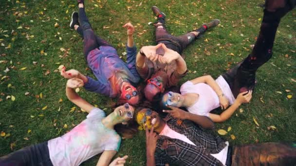 Top view of cheerful men and women in sunglasses with painted faces and clothes lying on meadow, looking at camera, smiling and moving hands. Nature and Holi concept. — Stock Video