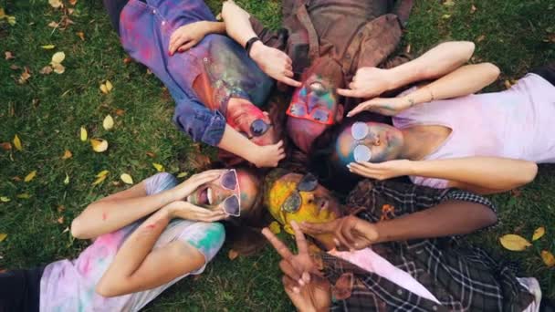 Slow motion top view of playful girls and guys with coloured faces lying on grass, posing and looking at camera, moving hands and smiling showing thumbs-up. — Stock Video