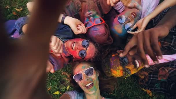 Slow motion synvinkel skott av sorglös studerande med färgade ansikten inspelning video tar selfie liggande på gräset tittar kameran och poserar rörliga händer. — Stockvideo