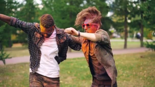 Two bearded guys with colorful faces covered with paint are dancing on lawn in park jumping and smiling enjoying party. Men with cool hairstyle are wearing sunglasses. — Stock Video