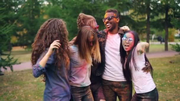 Retrato de jóvenes despreocupados bailando y saltando juntos abrazándose durante la fiesta en el césped en el parque, rostros, cabello y ropa están cubiertos de pintura . — Vídeos de Stock