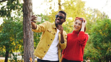 Afrika kökenli Amerikalı adam akıllı telefon tutarak ve parkta birlikte ayakta güneş gözlüğü ile poz kız arkadaşı ile selfie alıyor. Dostluk ve modern teknoloji kavramı.