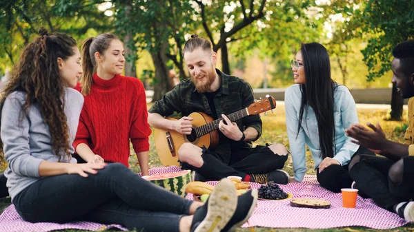 Радостный хипстер поет и играет на гитаре, сидя на одеяле в парке с друзьями и веселясь, люди хлопают в ладоши. Еда, музыка и развлечения . — стоковое фото