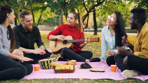 Charming young woman is playing the guitar sitting on blanket with friends on picnic, girls and guys are clapping hands and listening to music. Fun and nature concept.