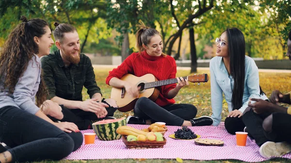 Bájos fiatal nő játszik a gitár meg a piknik takaró ült, a lányok és a fiúk taps kezek, és zenét hallgatni. Szórakozás és a természet koncepció. — Stock Fotó
