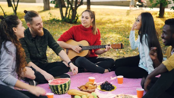 Красивая молодая леди играет на гитаре, пока ее друзья поют и слушают музыку, отдыхающую на клетке в парке. На одеяле видны фрукты и кондитерские изделия . — стоковое фото