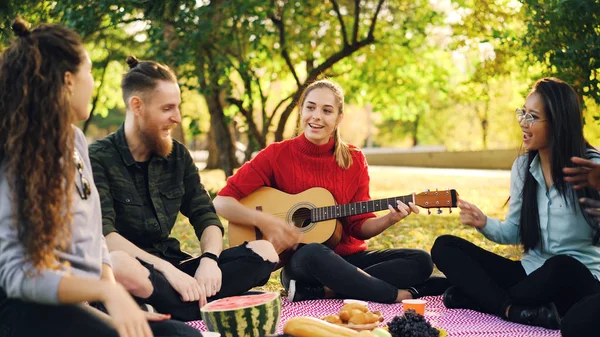Играющие молодые люди поют и двигают руками, когда красивая девушка играет на гитаре во время пикника в парке в солнечный осенний день. Концепция развлечений и музыки . — стоковое фото