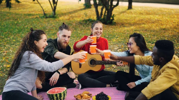 Веселая молодежь многонациональная группа звонят стаканы с напитками, то пить сидя на одеяле на траве во время осеннего пикника с гитарой и едой . — стоковое фото