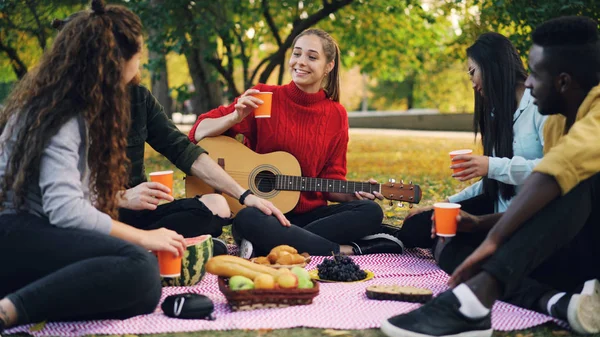 Giovani uomini e donne brindano e si accarezzano i bicchieri al picnic nel parco con la chitarra nelle calde giornate autunnali. Amicizia, bevande e attività ricreative concetto . — Foto Stock