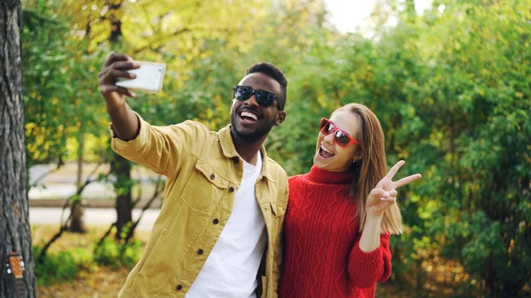 Schönes Paar afrikanisch-amerikanischer Mann und kaukasische Frau machen Selfie im Park mit Smartphone und posieren mit V-Zeichen und Zunge. Foto und Personenkonzept. — Stockfoto