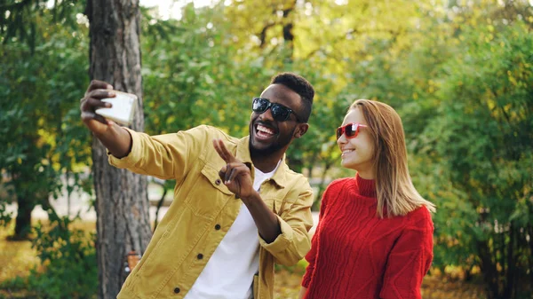 Beautiful couple African American man and Caucasian woman are taking selfie in park holding smartphone and posing showing v-sign and tongue. Photo and people concept.