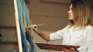 Beyaz gömlekli çekici genç kadın içinde modern studio çalışma fırça ve renk paleti tutan tuval üzerine yağlı boya resim deniz manzarası var. Sanat ve yaratıcılık kavramı.