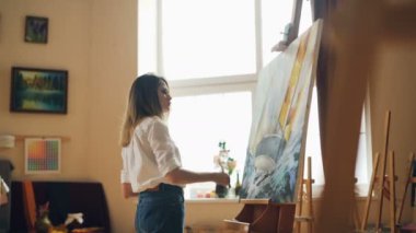 Meşgul genç kadın casual giyim şövale duran ve fırça ve palet tutan yağlıboya ile boyama. Hobi, sanat ve yaratıcı insanlar kavramı.