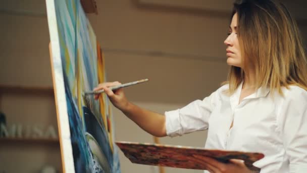 Atrakcyjna młoda kobieta w białej koszuli jest seascape malarstwo olej na płótnie, trzymając pędzla i kolor paleta praca w nowoczesnych apartamentów typu studio. Koncepcja sztuki i kreatywności. — Wideo stockowe
