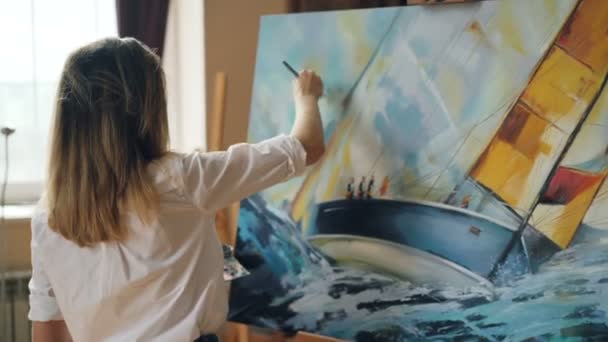Chica atractiva artista está ocupado pintando paisaje marino sobre lienzo utilizando paleta de colores, pincel y lienzo en caballete de trabajo en bonito estudio. Personas con talento y concepto de trabajo . — Vídeos de Stock