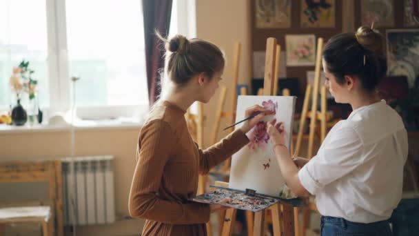 Studentka umělecké školy je malba květin s olejovými barvami, zatímco její učitel hezká mladá žena dává její Rady ukazuje na obrázek. — Stock video