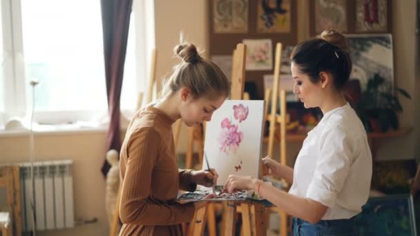 Mujer bonita profesora de pintura está enseñando a la joven a mezclar pinturas en la paleta creando un hermoso color durante la clase de arte en un bonito estudio moderno. Concepto de personas y educación . — Vídeos de Stock