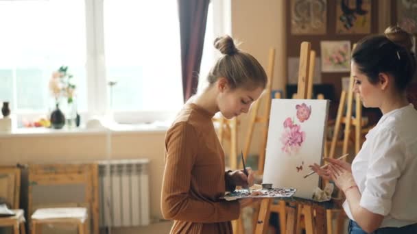 Jonge vrouwen kunst leraar en leerling ondervindt les op school van de schilderkunst, meisje is het mengen van kleuren op palet terwijl meester is het geven van advies gesprek met de leerling. — Stockvideo