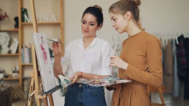 Female art teacher is explaining new information to student at painting lesson in studio pointing at picture and talking, girls are laughing having good time. — Stock Video