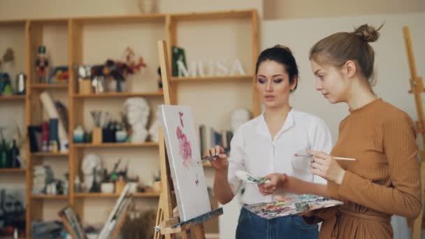 Experienced painting teacher is talking to her female student standing near picture on easel and discussing artwork. Shelves with tools are visible. — Stock Video