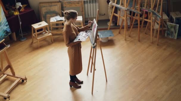 Vista de alto ângulo de pintora jovem trabalhando em estúdio em pé na frente de cavalete e pintura segurando escova e paleta. Móveis de madeira, obras de arte e ferramentas são visíveis . — Vídeo de Stock