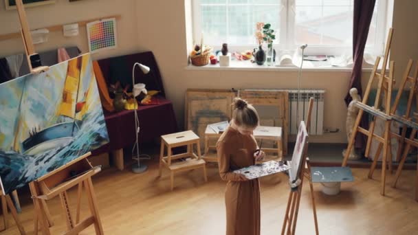 Creative person is painting in art studio using oil paints on palette and paintbrush, girl is concentrated on work. Young woman is wearing casual dress. — Stock Video