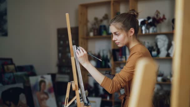 Jovencita guapa se centra en la pintura sosteniendo el pincel y trabajando con la imagen en caballete de pie solo en el taller lleno de obras de arte y herramientas . — Vídeo de stock