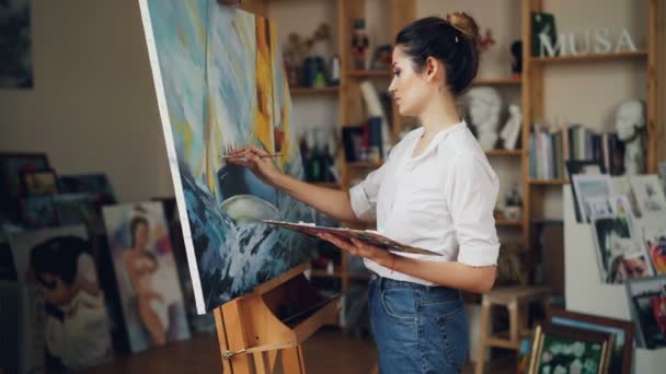 Allegro giovane artista donna sta dipingendo bel quadro paesaggio marino utilizzando vernici a olio poi guardando capolavoro e sorridente godendo il suo lavoro . — Video Stock