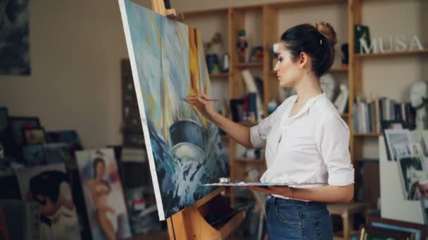 Glad female artist is painting picture depicting boat in sea then stepping back, looking at her work and smiling with satisfaction. Occupation and emotions concept. — Stock Video