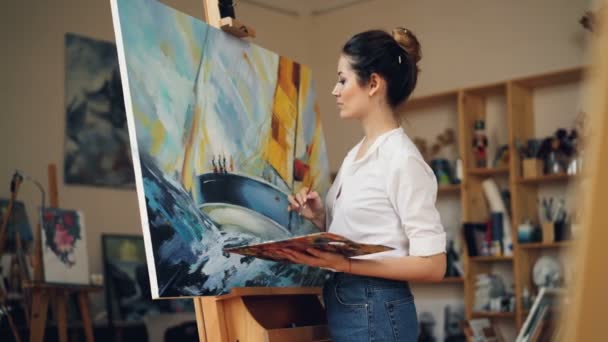 Skillful female painter is working in studio alone painting picture on easel using oil paints, palette and brush enjoying her occupation. People and work concept. — Stock Video