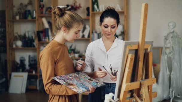 Friendly art teacher good-looking woman in casual clothing is teaching female student talking then giving her brush, girl is smiling and mixing paints on palette. — Stock Video