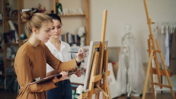 Cute teenage girl is learning painting from experienced teacher in art school in workroom with artworks and wooden easels. Artist is teaching talking and pointing at picture. — Stock Video