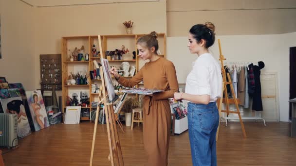 Slender young woman art student is painting working at picture and talking to her friendly teacher standing indoors in modern workshop. Fine arts and education concept. — Stock Video